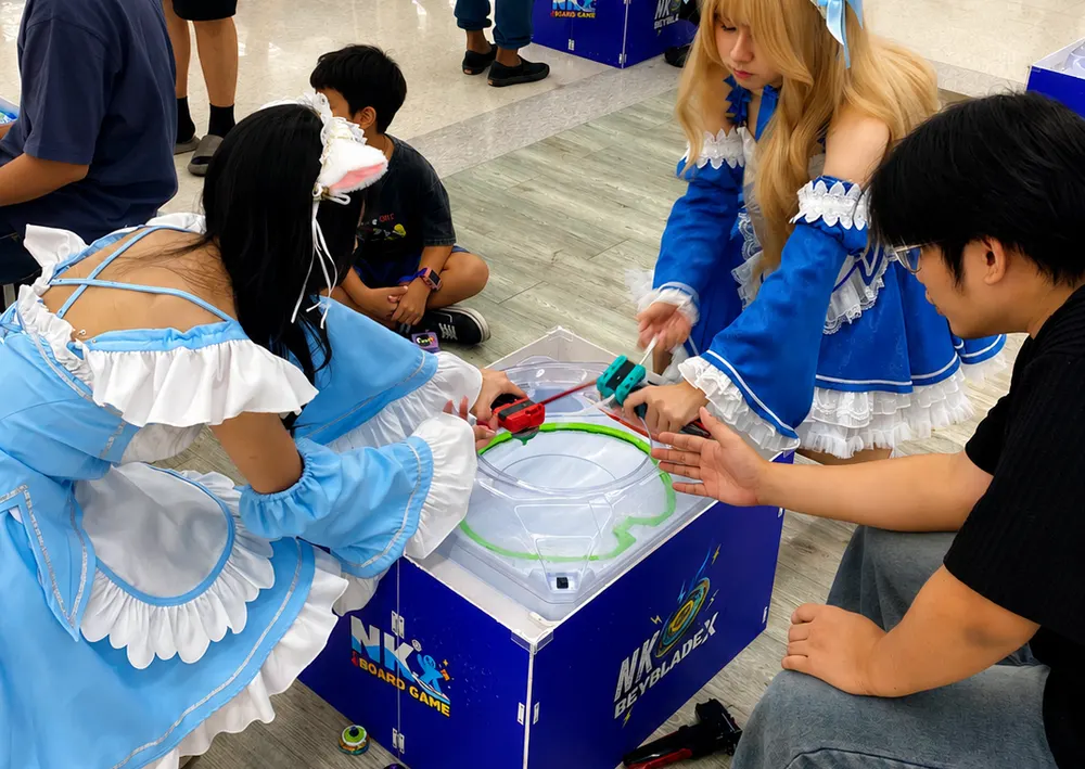 ผู้เล่นคอสเพลย์ชุดเมด 2 คน ชู้ต Beyblade X ลงสังเวียนสนามมาตรฐานของ NK Board Game ในกิจกรรม NK Beyblade X League